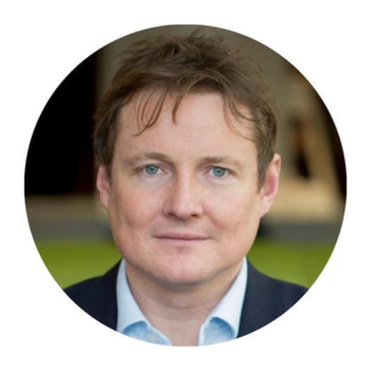 A circular headshot of Dr Tom Mitchell wearing a collared shirt and dark jacket, with a soft outdoor background featuring green and brown tones.